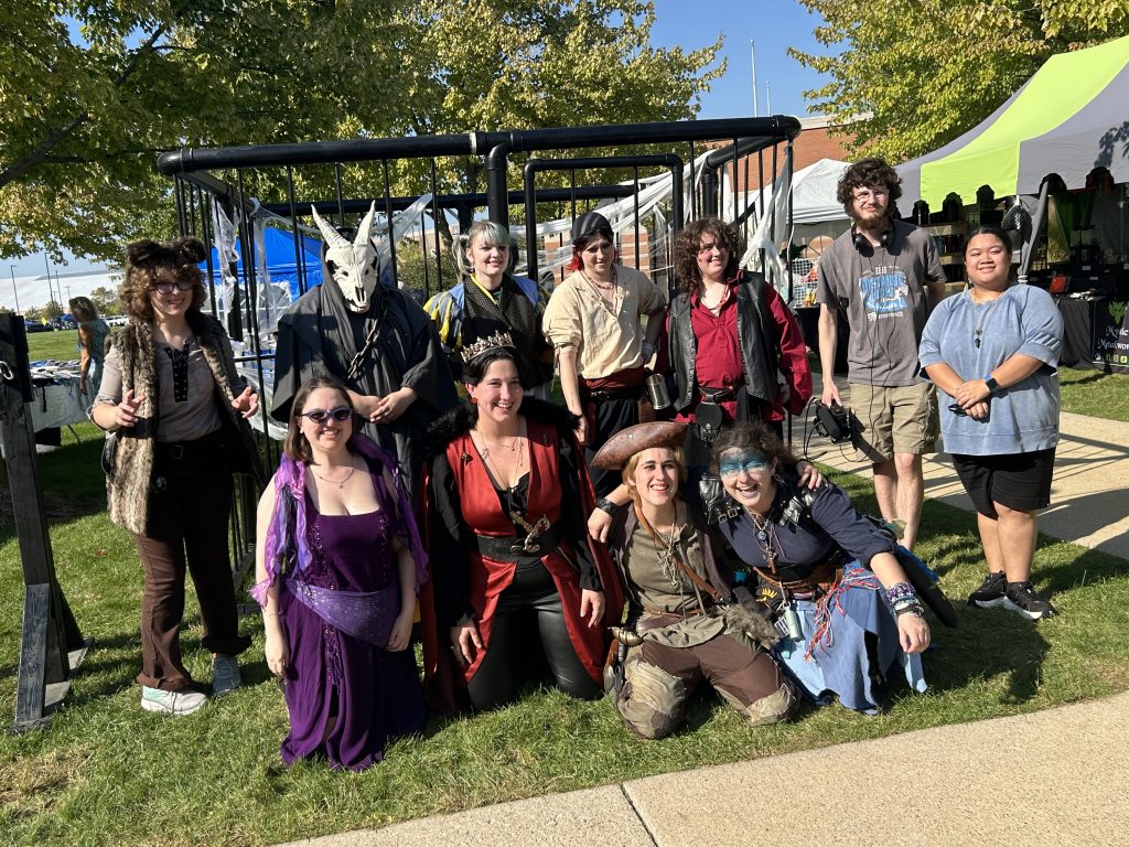 A group of college students and their faculty advisor posed in Renaissance costume and street clothes in front of a hand-made jail cell outside in the sunshine